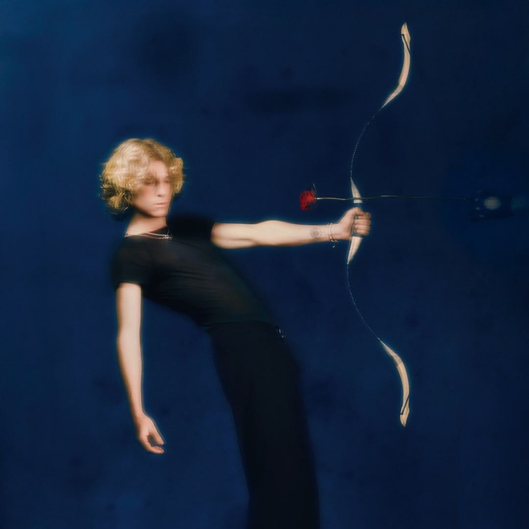 Noah Hill, sur fond bleu marine, tiens un arc donc la flèche pointe son coeur 