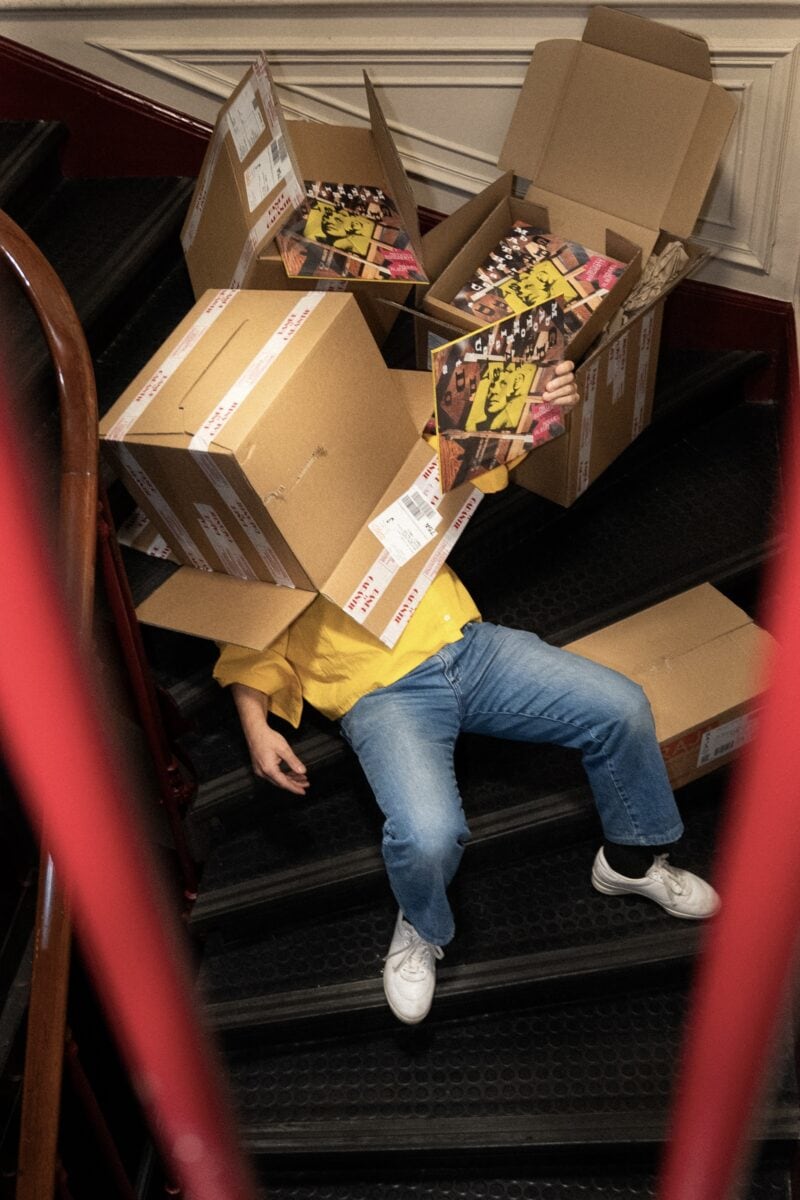 Photographie de Mathieu Boogaerts dans les escaliers de Tôt ou Tard, entouré d'exemplaires de son dernier album, Grand Piano. Sa tête disparait dans un carton. 