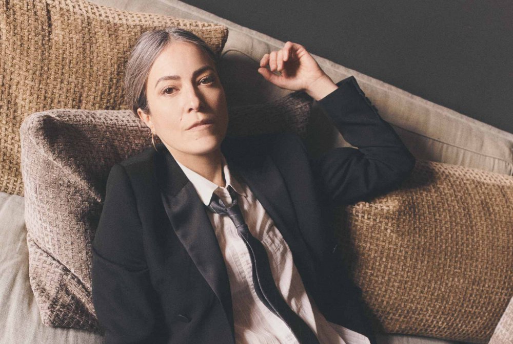 La photo montre la chanteuse et musicienne Keren Ann dans une pose décontractée. Vêtue d'un costume noir élégant, avec une chemise blanche et une cravate, elle semble à la fois sophistiquée et décontractée, se reposant sur des coussins en tissu beige. Son regard calme et légèrement pensif ajoute une touche d'intensité à l'image. La lumière douce met en valeur ses cheveux grisonnants et son expression sereine, soulignant une beauté intemporelle et une personnalité mystérieuse. Cette image reflète l'univers unique de Keren Ann, une artiste qui fusionne des éléments de la pop, du folk et du jazz dans ses créations musicales.