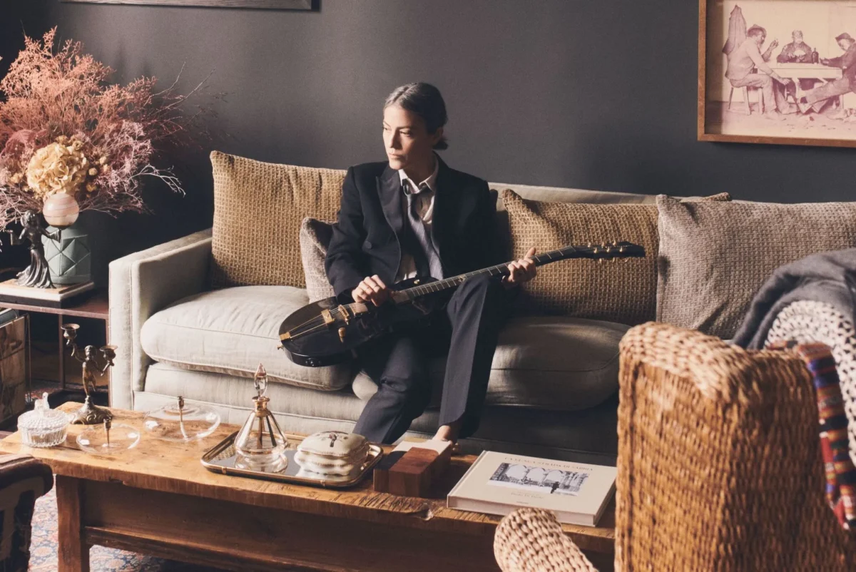 Dans un intérieur feutré aux teintes chaudes, une musicienne est assise sur un canapé en tissu clair, une guitare électrique posée délicatement sur les genoux. Vêtue d’un costume sombre à l’allure androgyne et élégante, elle adopte une posture calme et introspective, le regard tourné vers la gauche, comme absorbée par une pensée musicale. La lumière naturelle, douce et tamisée, souligne les textures du décor et crée une atmosphère intimiste.

Autour d’elle, le mobilier et les objets racontent une esthétique soignée : table basse en bois brut, verrerie ancienne, livres soigneusement disposés, fauteuil en rotin et compositions florales séchées. L’ensemble évoque un univers à la fois chaleureux, vintage et profondément artistique. Accrochée au mur, une photographie ancienne renforce ce sentiment de temps suspendu et de mémoire.

Cette image traduit un moment de pause et de création, où la musique semble prête à naître dans le silence. Elle incarne une sensibilité discrète, une élégance sans ostentation et une relation intime à l’instrument, à l’image d’un travail musical empreint de pudeur et de profondeur.