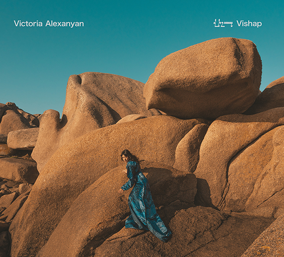 Une femme en robe bleue, Victoria Alexanyan, marche au pied d’imposants rochers dans un paysage désertique sous un ciel bleu clair. Elle est entre des pierres que l'on surnomme des Vishaps